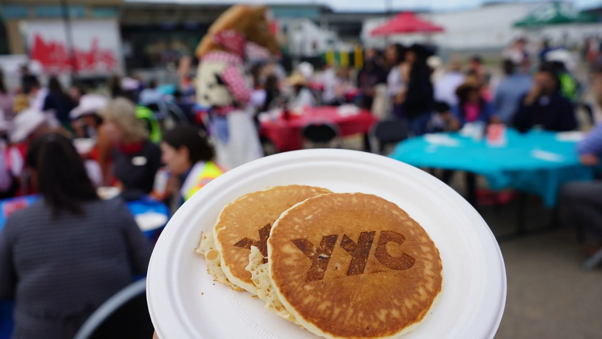 Close-up of a plate holding two golden-brown pancakes, each branded with the YYC logo, being held up at an outdoor community event with blurred crowds.