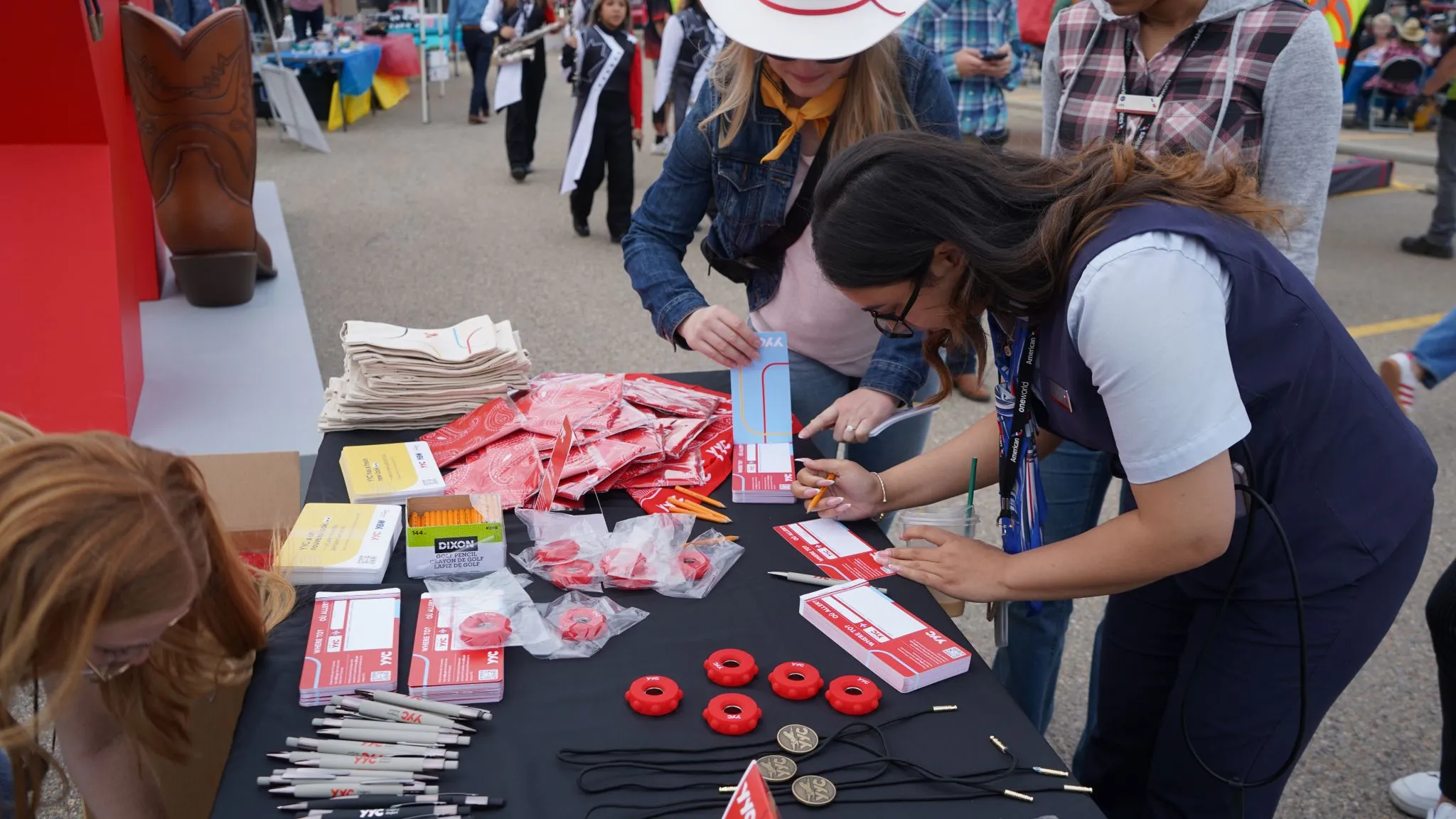 Several people, including one wearing a white cowboy hat, engaging at an outdoor booth with a black table covered in red and white branded promotional items like bandanas, pens, and keychains, with a large brown cowboy boot decoration in the background.